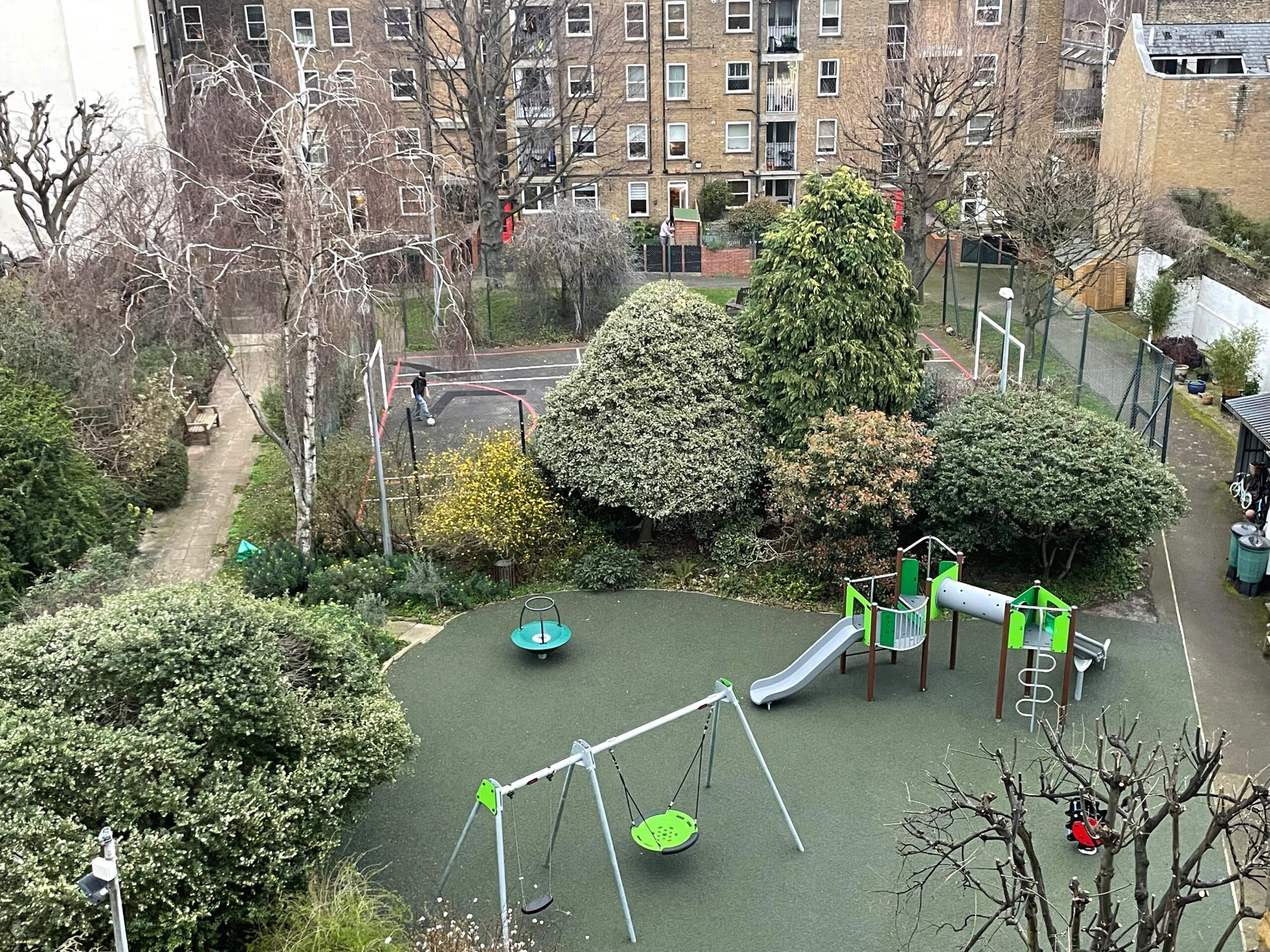 A bird's-eye view of a playground featuring swings, a slide, climbing structures, and a seating area. The playground is surrounded by trees and bushes. In the background, there is a fenced tennis court and a residential building.